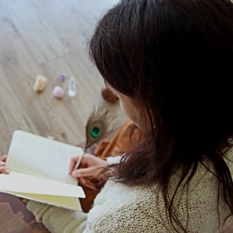 Persona escribiendo en un cuaderno, rodeada de piedras y plumas sobre el suelo.