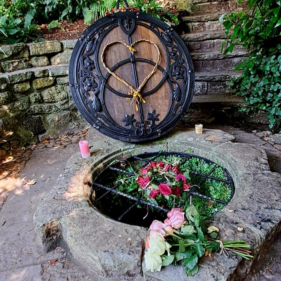 Limpieza energética hogar - Conciencia de corazon Escultura de piedra con una tapa decorada y flores en el borde de un pozo.