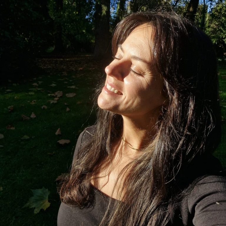 Mujer sonriendo al sol en un entorno natural con luz suave y hojas alrededor.