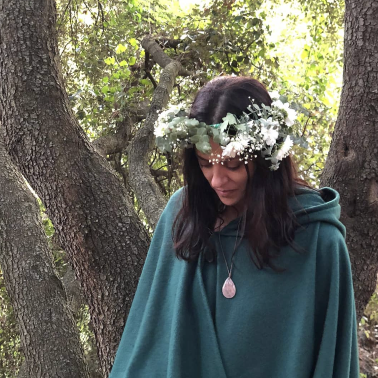 Mujer con capa verde y corona de flores, mirando hacia abajo en un entorno natural.